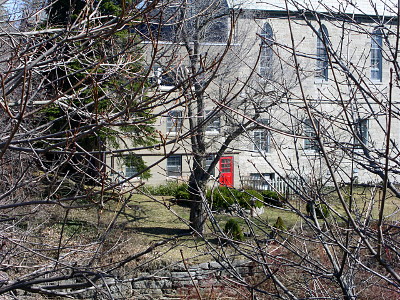 [The red door has nine windows in the top half and is a contrast to the light grey church which also has lots of windows. This image was taken through leafless trees whose branches cover the entire image.]